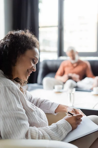 Smiling african american psychologist writing in notebook near blurred couple — Stock Photo