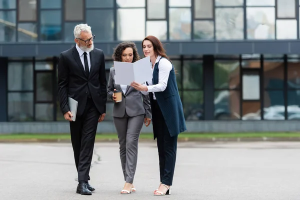 Real estate agent showing documents to interracial business colleagues near blurred building — Stock Photo