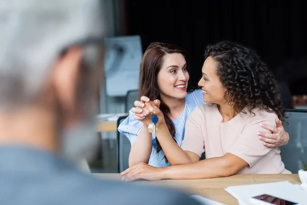 Happy lesbian woman hugging african american girlfriend holding key near blurred realtor — Stock Photo