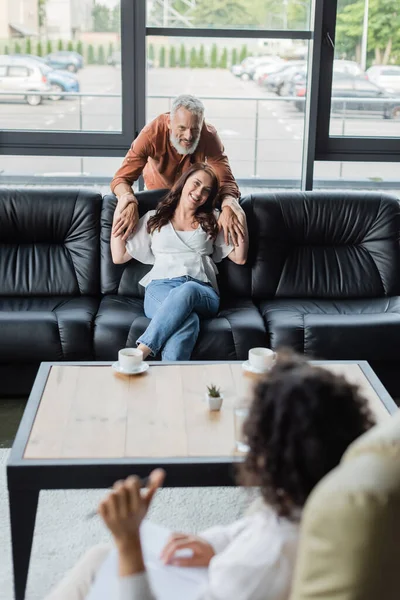 Felice uomo che abbraccia sorridente moglie seduta sul divano di fronte a sfocato psicologo afroamericano — Foto stock
