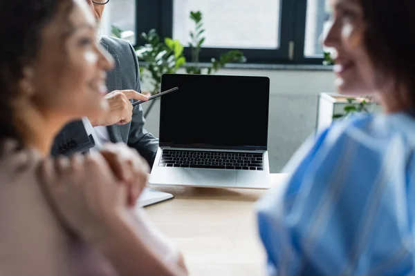 Real estate agent pointing at laptop with blank screen near interracial lesbian couple on blurred foreground — Stock Photo