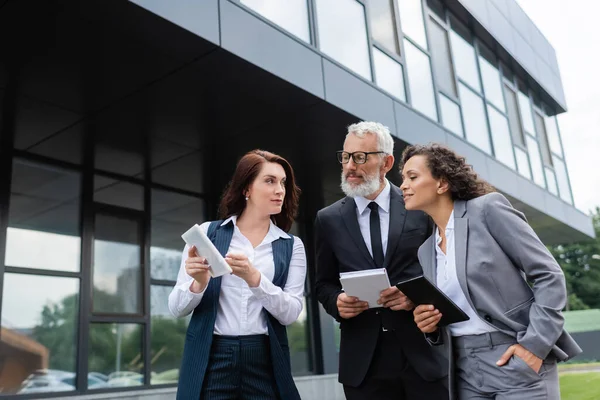 Real estate agent showing digital tablet to interracial business partners with notebooks near building — Stock Photo