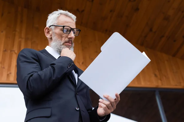 Homme d'affaires mûr réfléchi dans les lunettes de vue regardant des documents à l'extérieur — Photo de stock