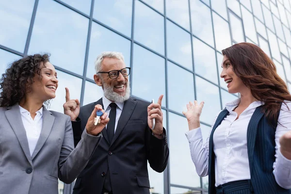 Emocionada mujer de negocios afroamericana sosteniendo la llave cerca de colega e inmobiliaria apuntando al edificio con fachada de vidrio - foto de stock