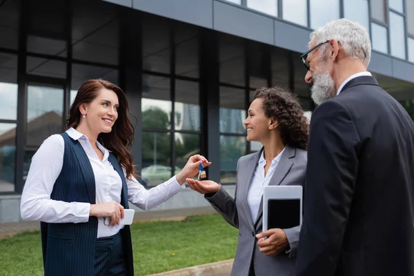 Lächelnder Makler übergibt Schlüssel an glückliche afrikanisch-amerikanische Geschäftsfrau mit digitalem Tablet und Notizbuch in der Nähe eines Kollegen — Stockfoto