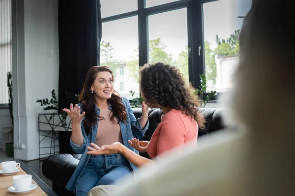 Excited interracial lesbian couple gesturing while talking in consulting room near blurred psychologist — Stock Photo