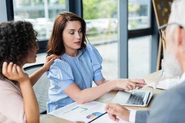 African american woman touching shoulder of lesbian girlfriend pointing with finger at laptop near blurred realtor — Stock Photo