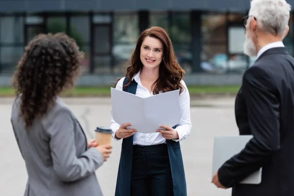 Real estate agent with folder smiling near blurred multiethnic business partners outdoors — Stock Photo