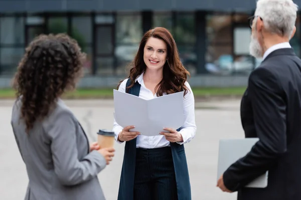 Agent immobilier avec dossier souriant près flous partenaires d'affaires multiethniques à l'extérieur — Photo de stock