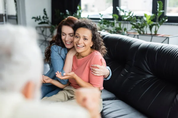 Feliz lesbiana mujer abrazando africano americano novia mientras sentado en sofá cerca psicólogo en borrosa primer plano - foto de stock