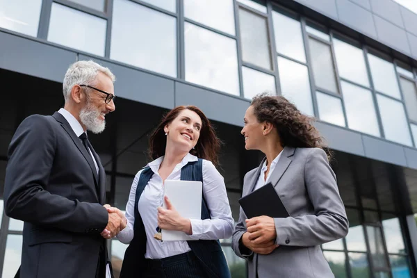 Feliz agente inmobiliario con tableta digital estrechando la mano con hombre de negocios de mediana edad cerca de su colega afroamericano - foto de stock