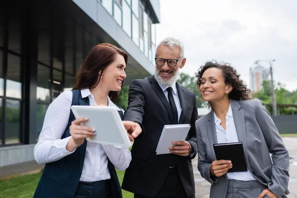 Alegre hombre de negocios de mediana edad apuntando a la tableta digital en manos de un agente inmobiliario cerca de un colega afroamericano - foto de stock