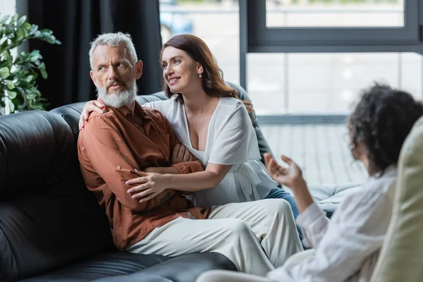 Donna sorridente che abbraccia un marito serio seduto sul divano con le braccia incrociate vicino allo psicologo afroamericano offuscato — Foto stock