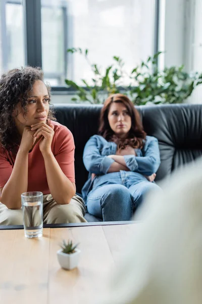 Depressed lesbian woman sitting on sofa with crossed arms near upset african american girlfriend and blurred psychologist — Stock Photo
