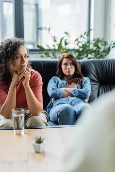 Déprimé lesbienne femme assis sur canapé avec les bras croisés près bouleversé afro-américaine copine et trouble psychologue — Photo de stock