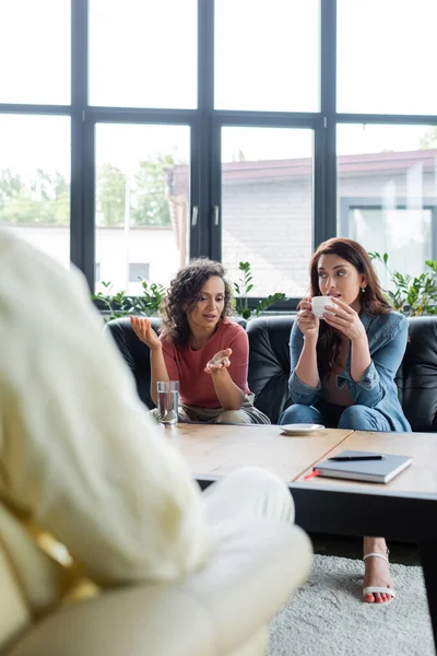 African american woman talking to lesbian girlfriend drinking coffee on couch during visit to blurred psychologist — Stock Photo