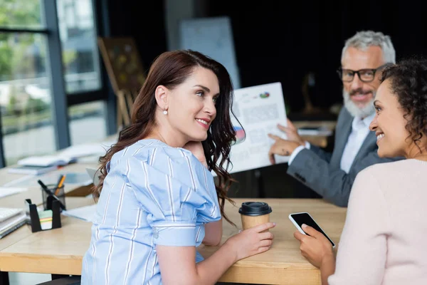 Pareja lesbiana interracial con café para llevar y teléfono inteligente mirándose cerca borrosa agente inmobiliario con gráficos hipotecarios - foto de stock