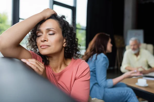 Depressed african american woman sitting with closed eyes near girlfriend and psychologist on blurred background — Stock Photo