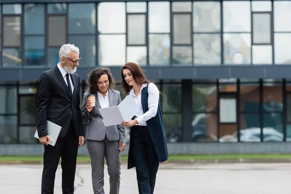 Real estate broker showing documents to multiethnic business partners near blurred building — Stock Photo