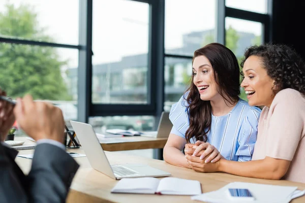 Joyful interracial lesbians holding hands while looking at laptop near blurred realtor — Stock Photo