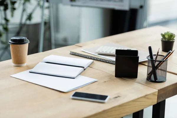 Telefono cellulare con schermo bianco vicino a notebook vuoto e tazza di carta sulla scrivania in ufficio — Foto stock