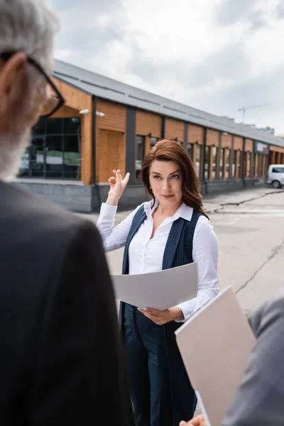 Agente de bienes raíces mirando a los socios de negocios borrosa mientras que apunta a la construcción al aire libre - foto de stock