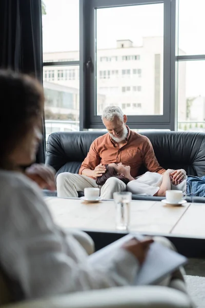 Smiling woman lying on couch near happy husband and blurred african american psychologist — Stock Photo