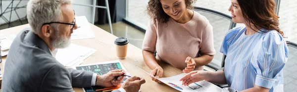 Smiling african american woman looking at documents with graphs near lesbian girlfriend and broker, banner — Stock Photo
