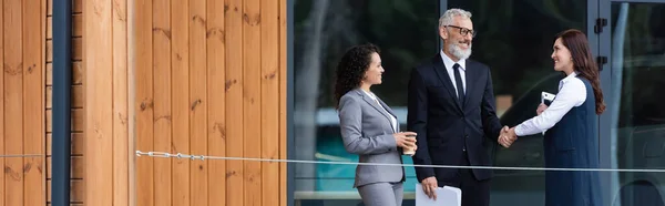 Happy businessman shaking hands with realtor near african american colleague and building outdoors, banner — Stock Photo