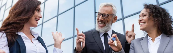 Happy african american businesswoman holding key near excited colleague and realtor pointing at building with glass facade, banner — Stock Photo