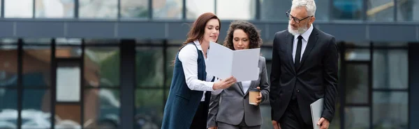 Agente inmobiliario mostrando documentos a socios comerciales interracial cerca de difuminado edificio al aire libre, pancarta - foto de stock