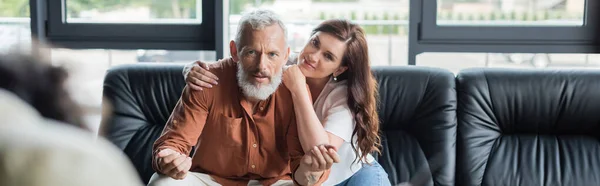 Smiling woman hugging middle aged husband gesturing while talking to blurred psychologist, banner — Stock Photo