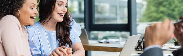 Aufgeregtes Paar interrassischer lesbischer Frauen, die Händchen halten, während sie auf Laptop in der Nähe des verschwommenen Maklers schauen, Banner — Stockfoto