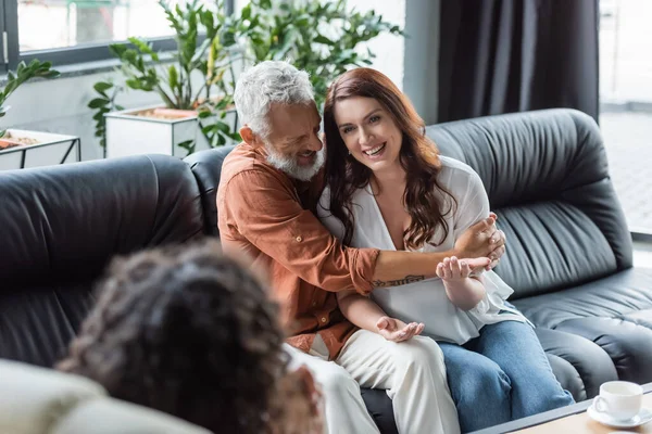 Homem alegre abraçando esposa sorridente enquanto sentado no sofá perto de psicólogo em primeiro plano borrado — Fotografia de Stock