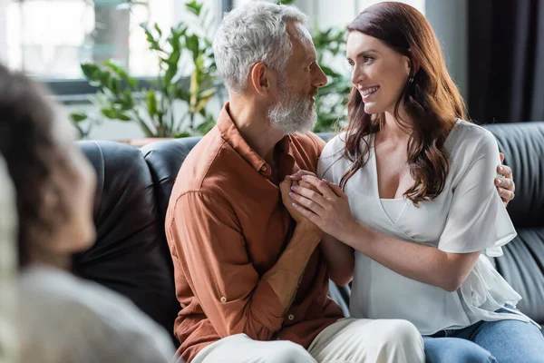 Feliz pareja cogida de la mano y mirándose mientras está sentada cerca borrosa afroamericana psicóloga en la sala de consulta - foto de stock