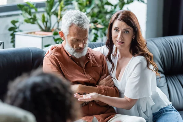 Donna preoccupata che parla con psicologo offuscata mentre abbraccia marito depresso seduto con le braccia incrociate — Foto stock