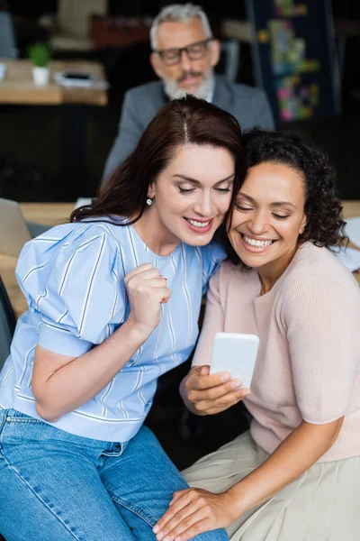 Alegre multiétnico casal do mesmo sexo olhando para o telefone móvel perto de corretor de imóveis de meia idade no fundo borrado — Fotografia de Stock