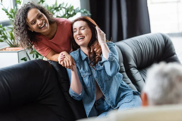 Aufgeregtes lesbisches Paar hält Händchen und lacht in der Nähe eines verschwommenen Psychologen während der Beratung — Stockfoto