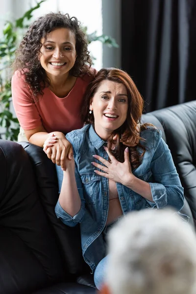 Excited and grateful lesbian woman touching chest while holding hand of african american girlfriend near blurred psychologist — Stock Photo