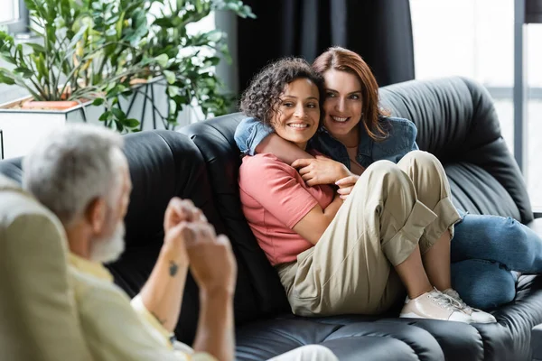 Cheerful couple of interracial lesbian women embracing on sofa during psychological consultation — Stock Photo