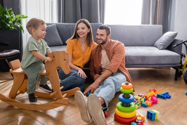 Cheerful parents looking at son on rocking horse in living room — Stock Photo