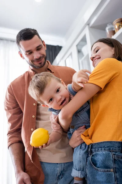 Sonriente madre sosteniendo hijo pequeño con limón cerca del marido en casa - foto de stock