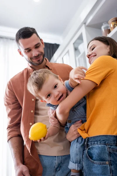 Mère souriante tenant tout-petit fils avec du citron près du mari à la maison — Photo de stock