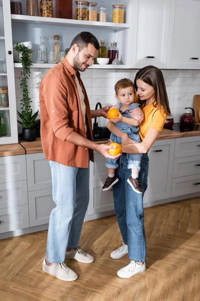 Sonrientes padres sosteniendo naranjas cerca de hijo en la cocina - foto de stock