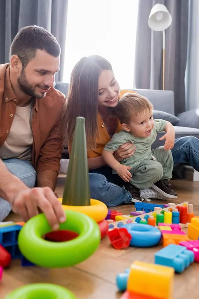 Sonrientes padres sentados cerca de juguetes y niños en la sala de estar - foto de stock