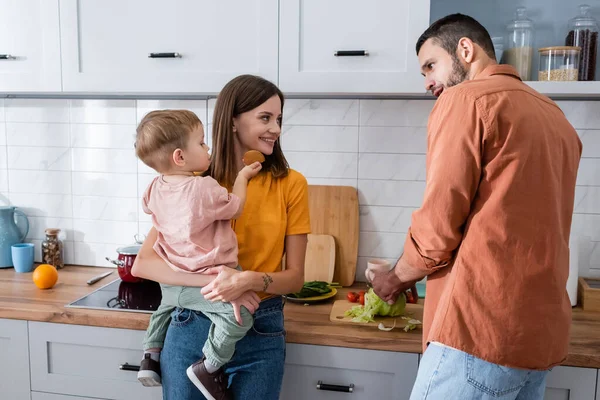 Mère souriante tenant enfant tandis que le mari coupe le chou dans la cuisine — Photo de stock