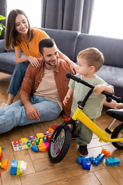 Niño pequeño de pie cerca de la bicicleta y padres positivos en la sala de estar - foto de stock