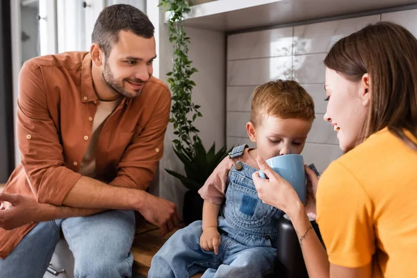 Smiling mom holding cup near son and husband in kitchen — Stock Photo