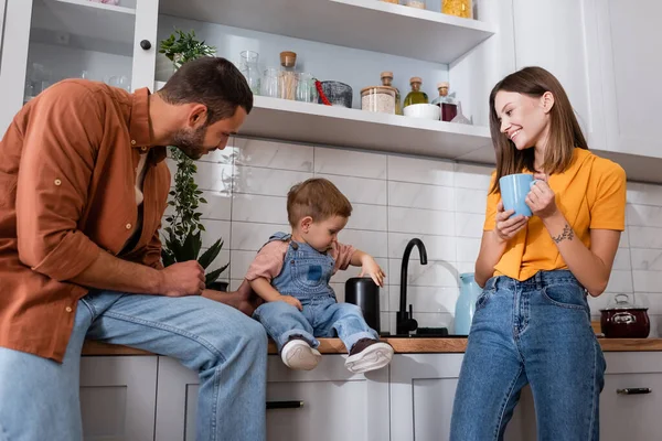 Frau mit Tasse sieht Kleinkind-Sohn und Ehemann in Küche — Stockfoto