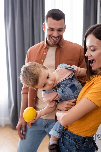 Aufgeregte Frau hält Kleinkind mit Zitrone neben verschwommenem Mann zu Hause — Stockfoto